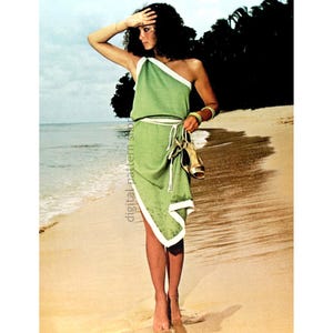 May include: A woman wearing a green one-shoulder dress with a white trim and a white belt. She is standing on a sandy beach with the ocean in the background.