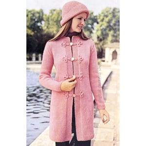 May include: A pink knitted cardigan with a high collar and decorative buttons. The cardigan has a long, fitted silhouette and is shown with a matching pink hat.