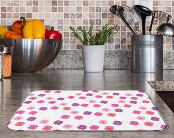 May include: A white kitchen counter with a pink and purple floral patterned dish drying mat. The mat is on a gray countertop with a stainless steel bowl filled with vegetables and a potted rosemary plant.
