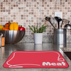 May include: A red rectangular kitchen mat with a white border and the word "Meat" in white block letters. The mat is on a countertop with a stainless steel bowl of vegetables, a potted herb, and kitchen utensils in the background.