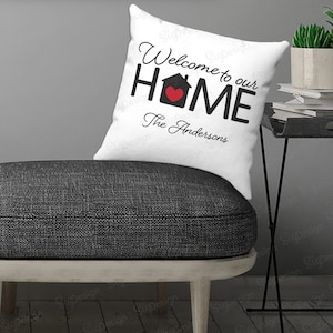 May include: White throw pillow with black text that reads "Welcome to our HOME" with a black house icon and a red heart inside. The text "The Andersons" is written in cursive below the house icon.