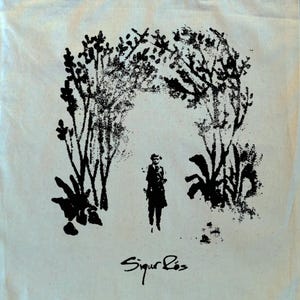 May include: Cream-colored tote bag featuring a black illustration of a person standing beneath an archway of trees and foliage. The text "Sigur R&oacute;s" is written in cursive at the bottom of the image.