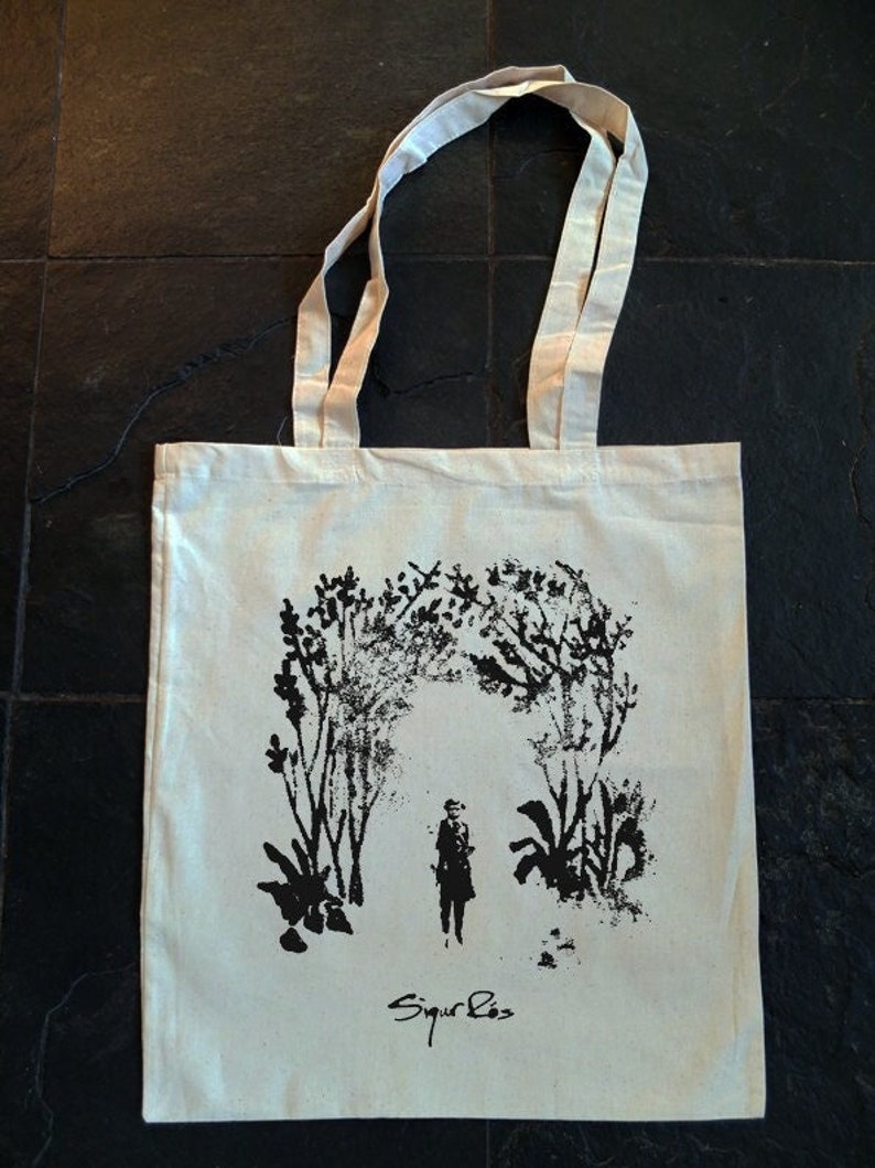 May include: Cream-colored tote bag with a black illustration of a person standing under an archway of trees. The text "Sigur Ros" is printed in black at the bottom. The bag has two cream-colored handles.