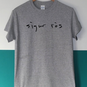 May include: A heather gray t-shirt with the handwritten text "sigur r&oacute;s" in black. The shirt is hanging on a wooden hanger against a white and teal background. The Gildan brand label is visible.