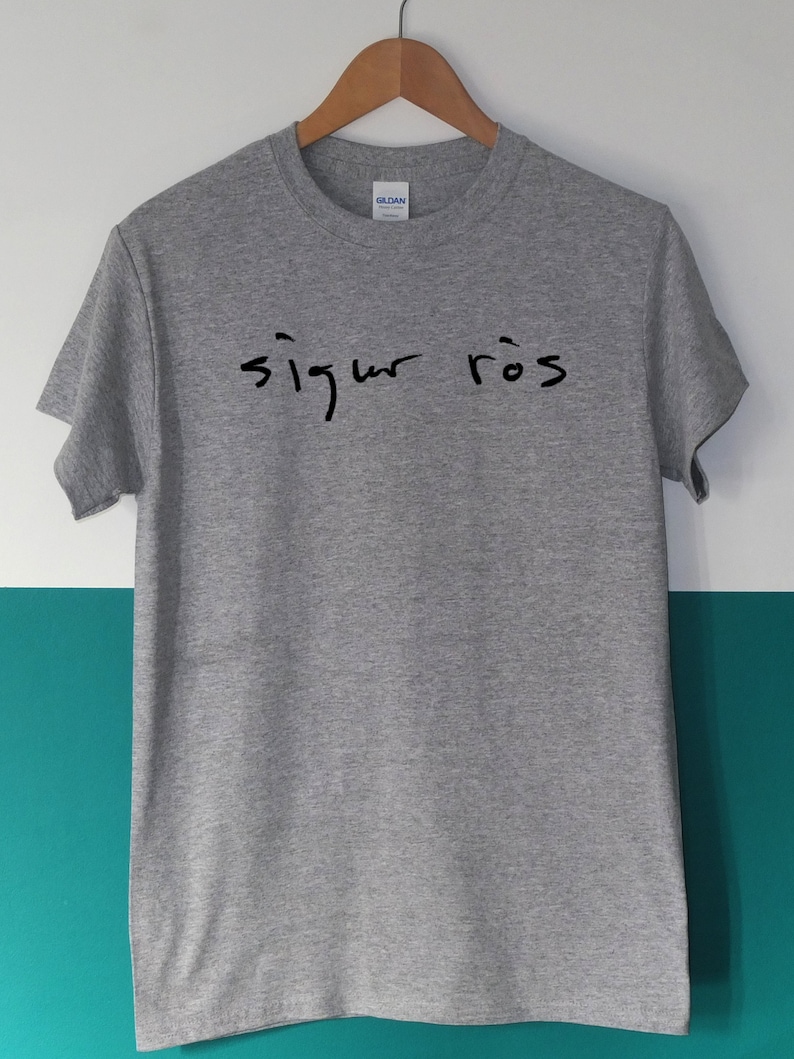 May include: A heather gray t-shirt with the handwritten text "sigur r&oacute;s" in black. The shirt is hanging on a wooden hanger against a white and teal background. The Gildan brand label is visible.