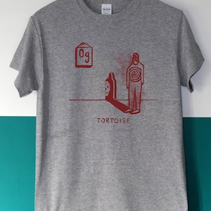 May include: A heather gray t-shirt with a red graphic design. The design includes a figure with a target on its chest, a shadow, and the word "TORTOISE". The shirt also features the text "09" in a box.