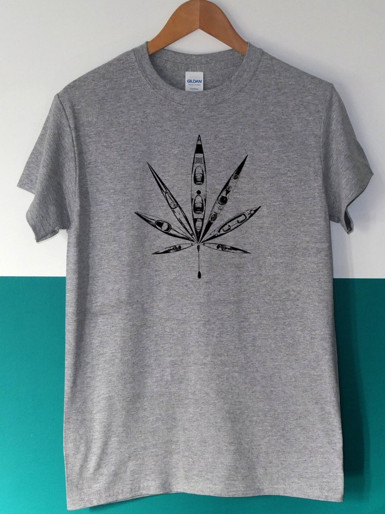 May include: A gray t-shirt featuring a black graphic of a cannabis leaf. The leaf design is filled with kayak illustrations. The shirt is hanging on a wooden hanger against a white and teal background.