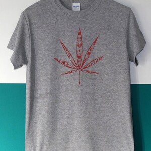 May include: A heather gray t-shirt featuring a red leaf design. The leaf is composed of several kayak illustrations, creating a unique and eye-catching graphic. The shirt is displayed on a wooden hanger.