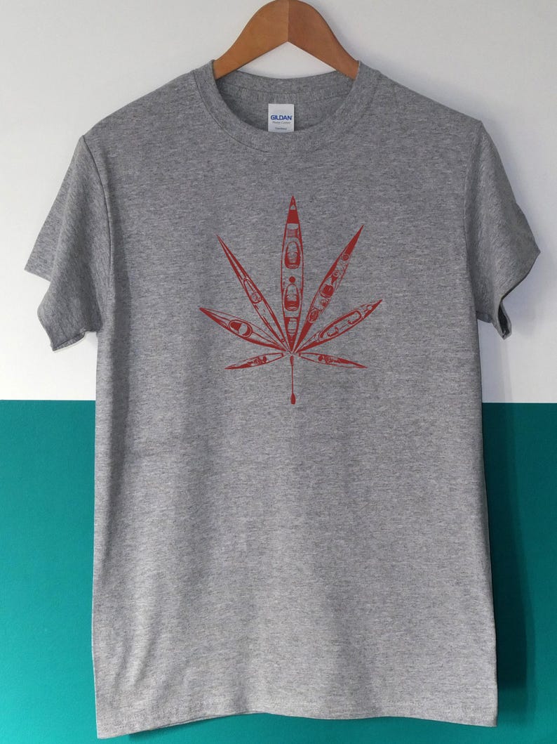 May include: A heather gray t-shirt featuring a red leaf design. The leaf is composed of several kayak illustrations, creating a unique and eye-catching graphic. The shirt is displayed on a wooden hanger.