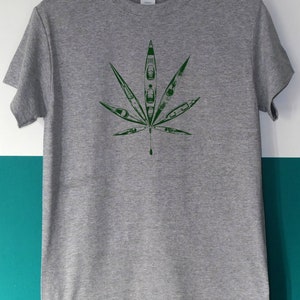 May include: A gray t-shirt featuring a green leaf design. The leaf is composed of kayak illustrations. The shirt has a crew neck and short sleeves. The brand name "GILDAN" is visible on the tag.
