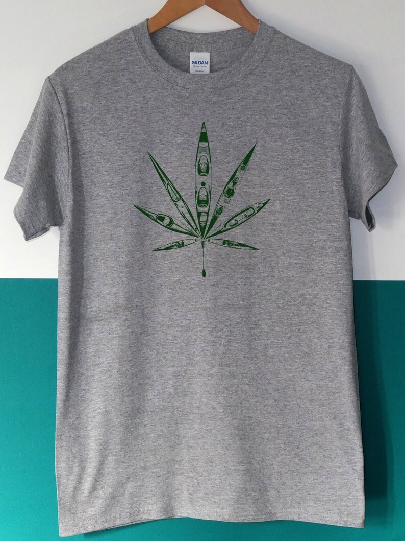May include: A gray t-shirt featuring a green leaf design. The leaf is composed of kayak illustrations. The shirt has a crew neck and short sleeves. The brand name "GILDAN" is visible on the tag.