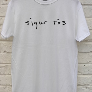 May include: White Gildan t-shirt with the handwritten text "sigur r&oacute;s" in black across the front. The shirt is hanging on a wooden hanger against a white brick wall.