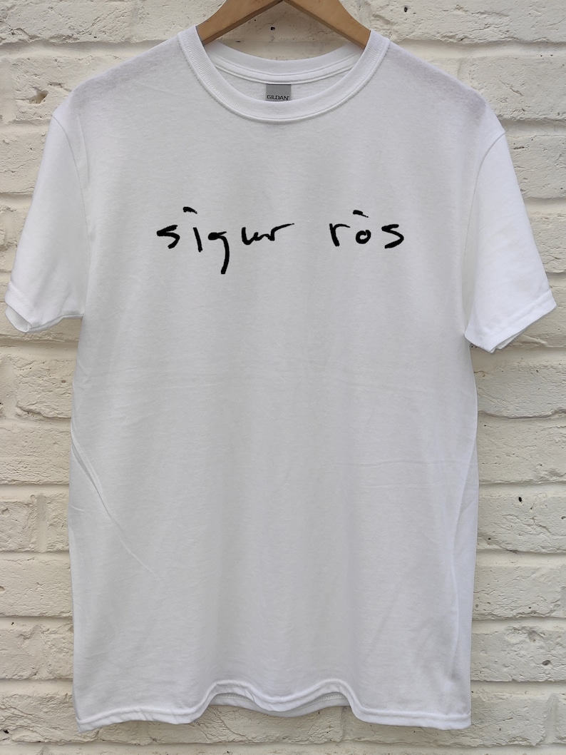 May include: White Gildan t-shirt with the handwritten text "sigur r&oacute;s" in black across the front. The shirt is hanging on a wooden hanger against a white brick wall.