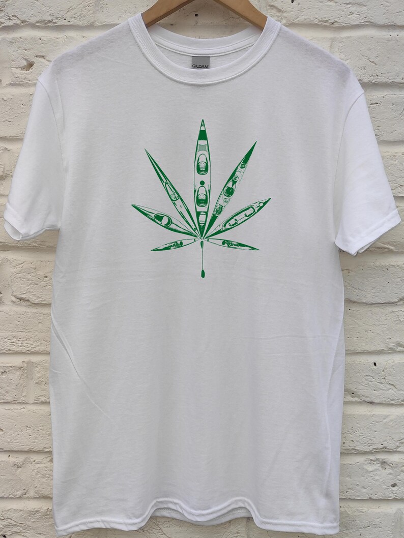 May include: White t-shirt featuring a green graphic of a cannabis leaf. The leaf design is filled with kayak illustrations. The shirt is made of a soft material and has a crew neck.