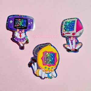 May include: Three holographic stickers featuring a girl with a purple Game Boy Color, a girl with a pink and white computer, and a girl with a yellow Tamagotchi. Each sticker has a glittery finish.
