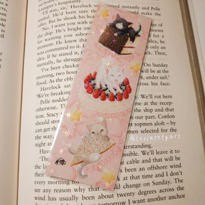 May include: A pink bookmark with three cat illustrations. One cat is on a chocolate cake, another on a cake with berries, and the third with fairy bread. Text includes "Lamington cat", "Pavlova cat", and "fairy bread cat". The bookmark is designed for use in books.