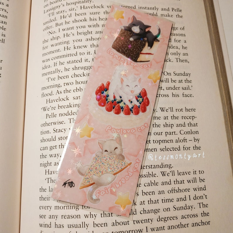 May include: A pink bookmark with three cat illustrations. One cat is on a chocolate cake, another on a cake with berries, and the third with fairy bread. Text includes "Lamington cat", "Pavlova cat", and "fairy bread cat". The bookmark is designed for use in books.