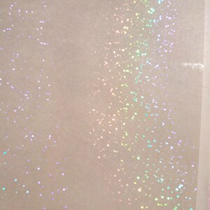 May include: Close-up of a white, textured surface with iridescent glitter scattered across it. The glitter reflects a spectrum of colors, including pink, green, and blue, creating a shimmering effect. The background is a soft, off-white color.