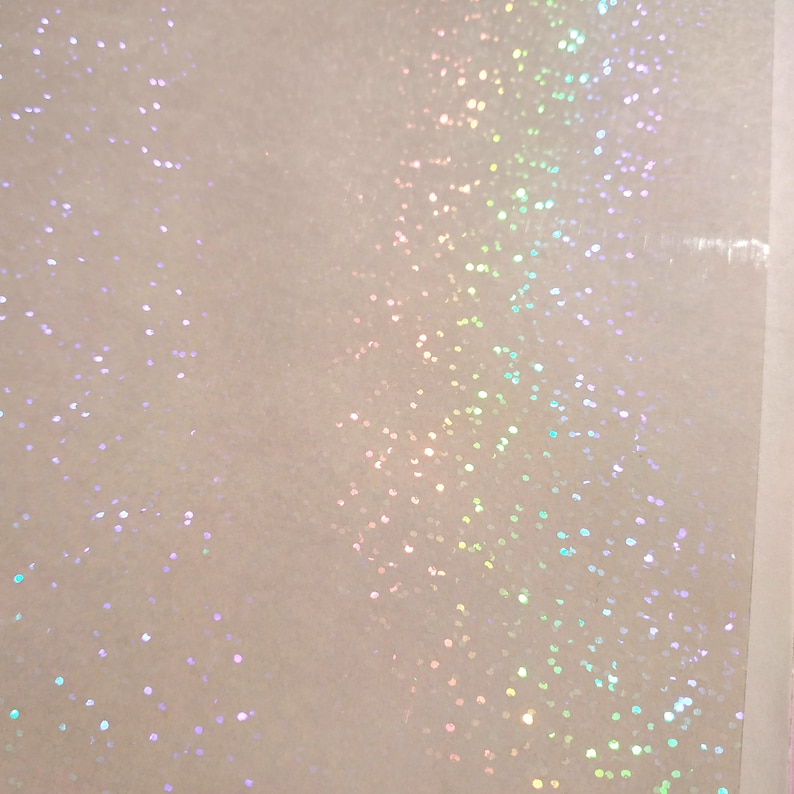 May include: Close-up of a white, textured surface with iridescent glitter scattered across it. The glitter reflects a spectrum of colors, including pink, green, and blue, creating a shimmering effect. The background is a soft, off-white color.