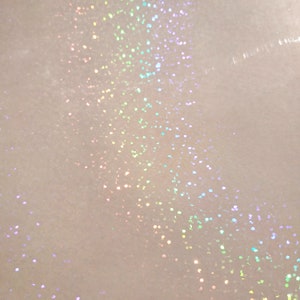 May include: Close-up of a white surface with scattered iridescent glitter. The glitter particles reflect a spectrum of colors, including pink, green, and blue, creating a shimmering effect. The background is a soft, off-white color.