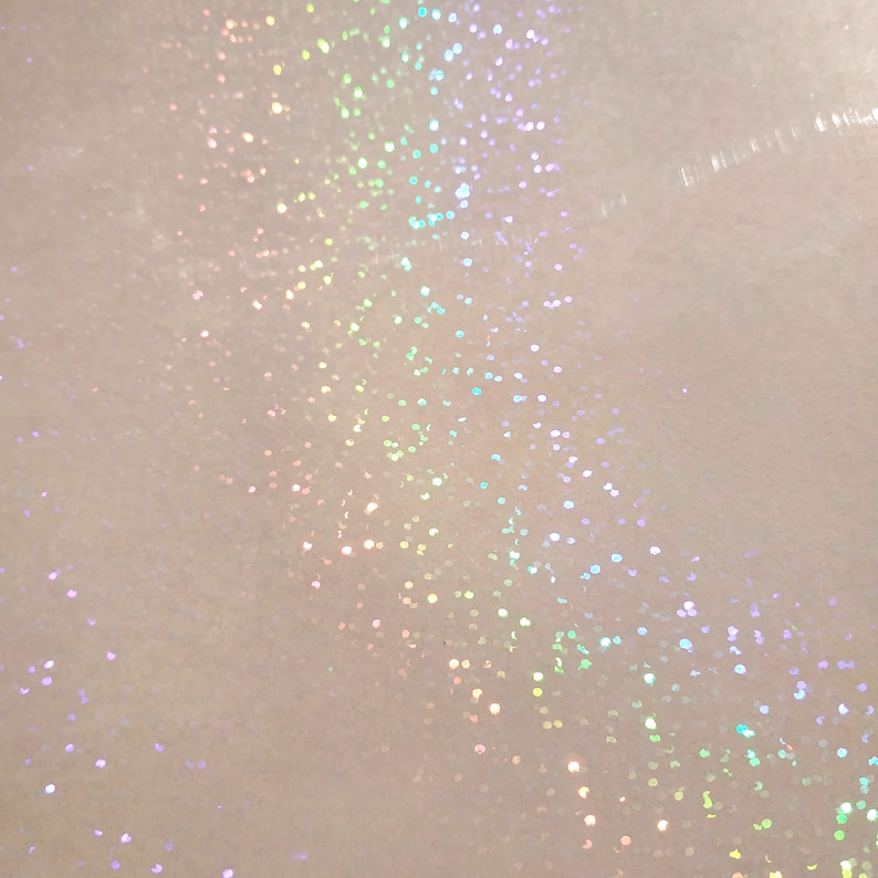 May include: Close-up of a white surface with scattered iridescent glitter. The glitter particles reflect a spectrum of colors, including pink, green, and blue, creating a shimmering effect. The background is a soft, off-white color.