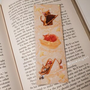 May include: A decorative bookmark with three cat illustrations. One cat is on toast, another on a pie, and the last in a sausage. The bookmark has a light yellow background with star accents and text.