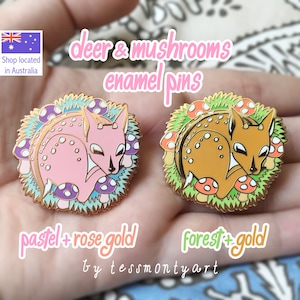 May include: Two enamel pins featuring deer and mushrooms. One pin is pastel pink with rose gold accents, the other is forest brown with gold accents. The pins are surrounded by a green and blue border, with the text "deer & mushrooms enamel pins".
