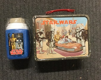 Puede incluir: Una fiambrera vintage de Star Wars con un asa roja y una escena de personajes de la película en la parte delantera. La fiambrera está hecha de metal y tiene un diseño marrón y negro. La fiambrera está al lado de un termo Star Wars azul y blanco.