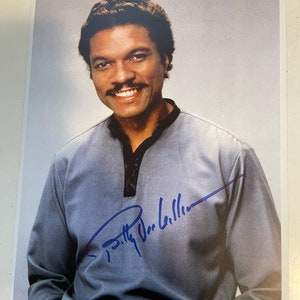 May include: A black and white photograph of a man with a moustache smiling. He is wearing a blue shirt and has signed his name in blue ink. The signature reads "Billy Dee Williams".