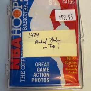 May include: A clear plastic case containing a blue and red NBA Hoops trading card. The card features a basketball player and the text "NBA Hoops, The Official Basketball Card." A yellow sticky note with handwritten text: "1989 Michael Jordan on Top." is also visible.