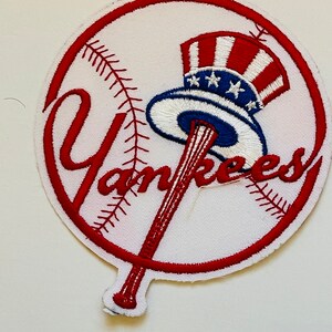 Puede incluir: Un parche bordado blanco con un círculo rojo alrededor de una pelota de béisbol con costuras rojas. La palabra "Yankees" está bordada en rojo con un bate de béisbol y un sombrero rojo, blanco y azul en el centro.