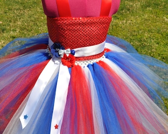 May include: Red, white, and blue tulle tutu with a red sequined bodice and a white satin sash. The tutu has red, white, and blue stars on the tulle.