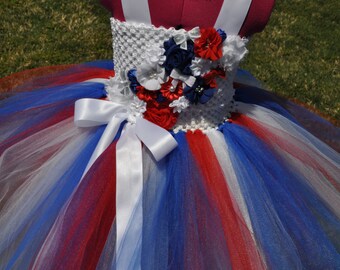 May include: A red, white, and blue tutu with a white crocheted bodice and a white satin bow. The bodice is decorated with red, white, and blue flowers.