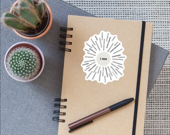 May include: A brown notebook with a spiral binding and a white sticker with the words "I AM" in the center surrounded by a circle of positive adjectives. A brown pen is lying on top of the notebook.
