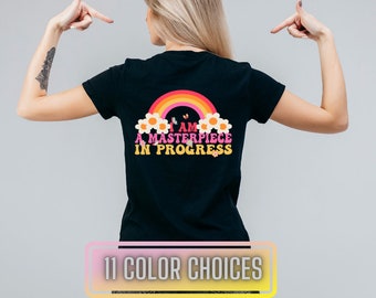 May include: Black t-shirt with a rainbow, flowers, and butterflies graphic on the back. The text on the graphic reads "I am a masterpiece in progress".
