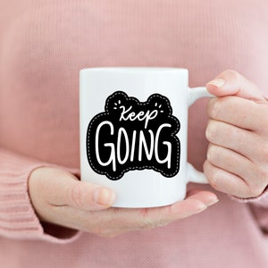 Puede incluir: Taza de cerámica blanca con un gráfico en blanco y negro que dice "Keep Going" en un estilo de escritura a mano.