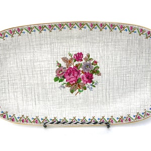 Könnte beinhalten: Ein weißes ovales Serviertablett mit goldfarbenem Rand und einem floralen Design in der Mitte. Das florale Design zeigt rosa, rote und violette Blumen mit grünen Blättern. Das Tablett hat ein gewebtes Muster auf der Oberfläche.