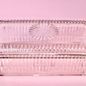 Puede incluir: Un plato de vidrio transparente rectangular con tapa. El plato presenta un diseño texturizado y acanalado en los lados y un patrón de rayos de sol en la tapa. Los bordes están detallados con un borde de cuentas. El plato es probablemente para servir o almacenar.