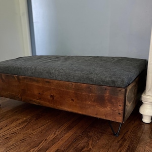 May include: A wooden storage bench with a gray fabric cushion and black hairpin legs. The bench is made of reclaimed wood and has a rustic look.