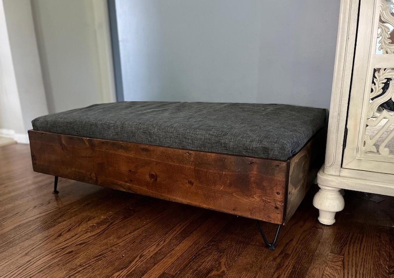 May include: A wooden storage bench with a gray fabric cushion and black hairpin legs. The bench is made of reclaimed wood and has a rustic look.