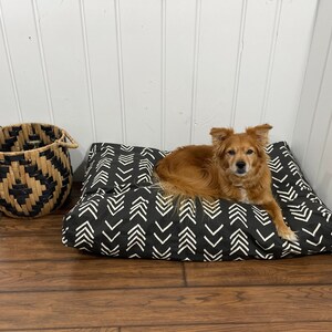 May include: A black and white patterned dog bed with a brown and white dog laying on it. The dog bed is on a wooden floor. There is a woven basket in the background.