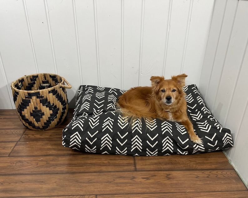 May include: A black and white patterned dog bed with a brown and white dog laying on it. The dog bed is on a wooden floor. There is a woven basket in the background.