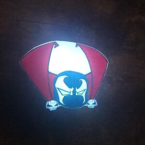 May include: A cartoon-style image of the Spawn character. The design features a blue head with white accents, a red and white cape, and two skull-shaped elements at the bottom. The background is dark.
