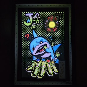 May include: A framed artwork featuring a cartoon shark with a menacing expression, holding a gauntlet with colorful gems. The artwork includes the text "Jeff The Land Shark" and question mark symbols, set against a dotted background.