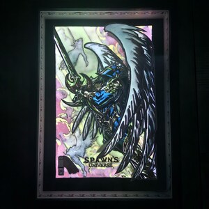 May include: Framed artwork featuring a detailed illustration of a winged warrior with a sword, set against a colorful background. The artwork is labeled "Spawn's Universe" and is encased in a decorative frame.