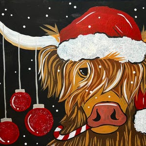 May include: A brown and white Highland cow wearing a red Santa hat with white trim and a candy cane in its mouth. The cow is surrounded by red Christmas ornaments hanging from strings.