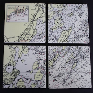 May include: A set of four square coasters featuring detailed nautical charts of the Gulf of Maine and Casco Bay. Each coaster displays cream-colored land masses, white water areas, black text labels, depth soundings, and red navigational markers.