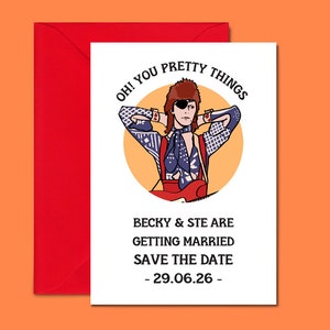 May include: A white card with a colorful illustration of a person with an eye patch and a guitar. The text on the card reads "OH! YOU PRETTY THINGS BECKY & STE ARE GETTING MARRIED SAVE THE DATE - 29.06.26 - "