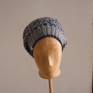 Può includere: Un berretto lavorato a maglia grigio e blu navy con bordo a coste e corona fantasia. Il cappello è esposto su una testa di manichino in legno. Il cappello ha un bordo blu scuro.