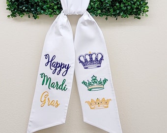 Może przedstawiać: Biała wstążka z haftem "Happy Mardi Gras" w kolorze złotym, zielonym i fioletowym. Na wstążce wyszyte są trzy złote, zielone i fioletowe korony.